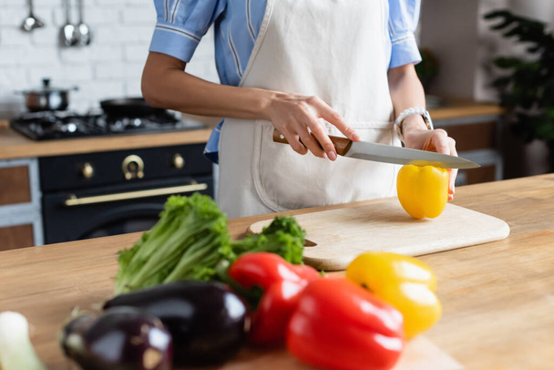 vista parcial de la mujer joven adulta cortando pimiento amarillo en la tabla de cortar en la cocina - Foto, imagen