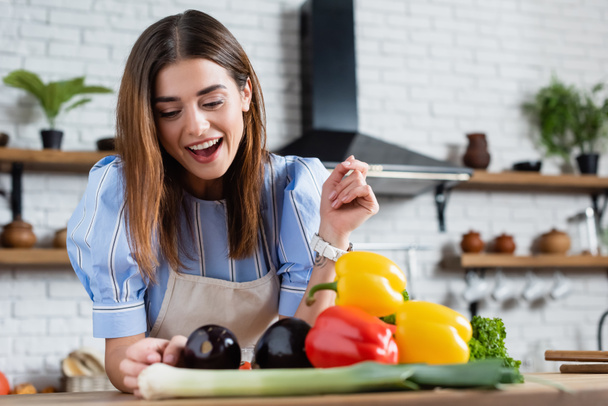 positive young adult woman looking at fresh vegetables on table in kitchen - Фото, изображение