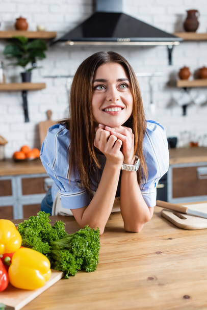young adult woman standing near table with vegetables and holding hands near face in kitchen - Foto, Imagen