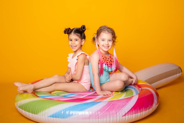 İki mutlu küçük kız mayolu renkli şişme yatakta oturuyor sarı arka planda lolipop. - Fotoğraf, Görsel