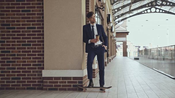 volledige lengte van zakenman in bril en pak houden koffie te gaan terwijl poseren in de buurt penny board in winkelcentrum  - Foto, afbeelding