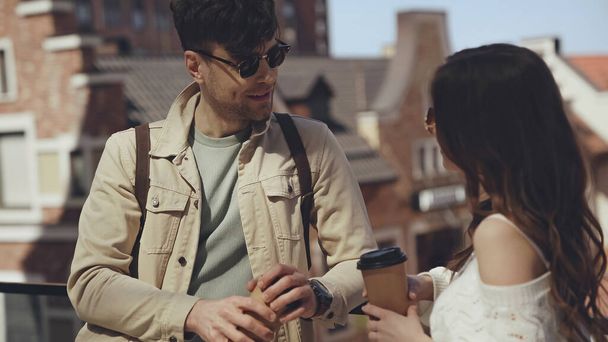 stylowa para w okularach przeciwsłonecznych trzymająca papierowe kubki i rozmawiająca na zewnątrz  - Zdjęcie, obraz