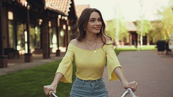 positive young woman in yellow blouse smiling while riding bicycle outside - Foto, Imagem