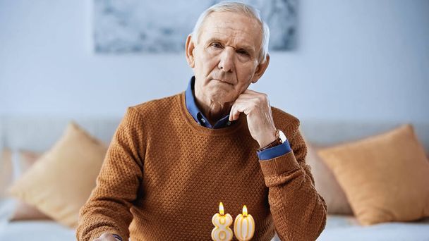 μοναχικός ηλικιωμένος που γιορτάζει τα γενέθλιά του μπροστά σε κεριά που καίγονται στο σαλόνι - Φωτογραφία, εικόνα