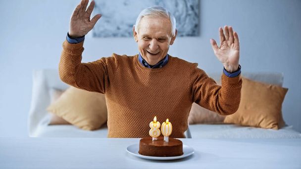 vreugdevolle oudere man viert verjaardag voor taart met brandende kaarsen in de woonkamer - Foto, afbeelding