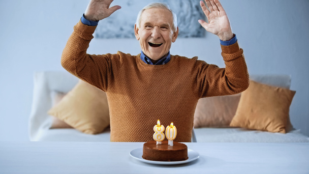 excited elderly man celebrating birthday in front of cake with burning candles in living room - Photo, Image