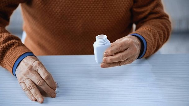 καλλιεργημένη άποψη του ανοιχτού βάζου ιατρικής στο χέρι του ηλικιωμένου άνδρα σε γκρι φόντο - Φωτογραφία, εικόνα
