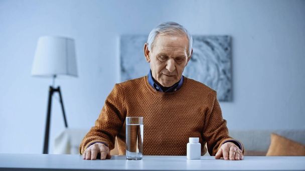 Älterer Mann sitzt neben Tisch mit Medikamentenglas und Wasserglas im Wohnzimmer - Foto, Bild