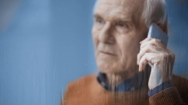 sad elderly man looking outside through rainy window and speaking on cellphone grey background behind rainy glass - Fotó, kép