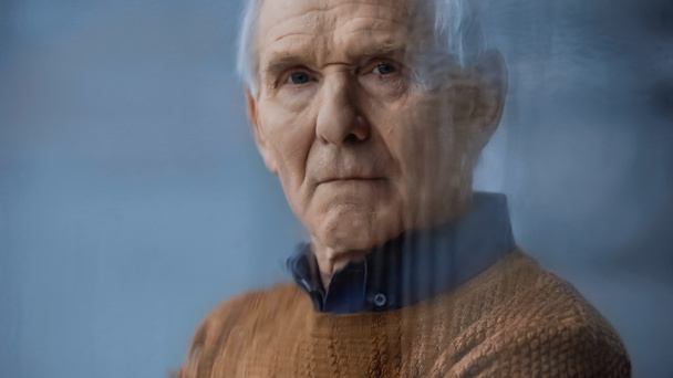 portrait d'un homme âgé regardant la caméra à travers une fenêtre pluvieuse sur fond gris derrière un verre pluvieux - Photo, image