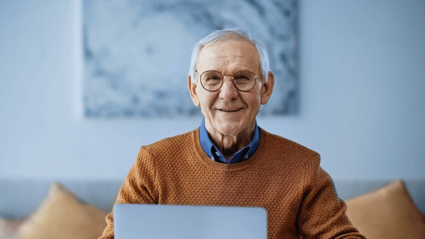 улыбающийся пожилой мужчина сидит дома с ноутбуком - Фото, изображение
