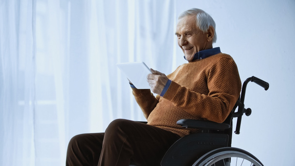smiling elderly man in wheelchair looking at tablet in hands at retirement home - Photo, Image