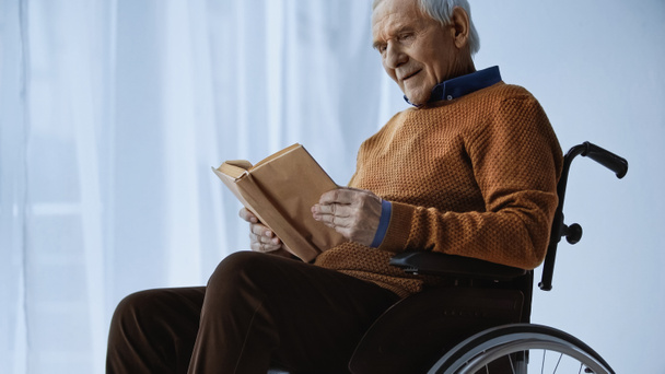 hombre mayor sentado en silla de ruedas y leyendo libro en casa de retiro - Foto, imagen