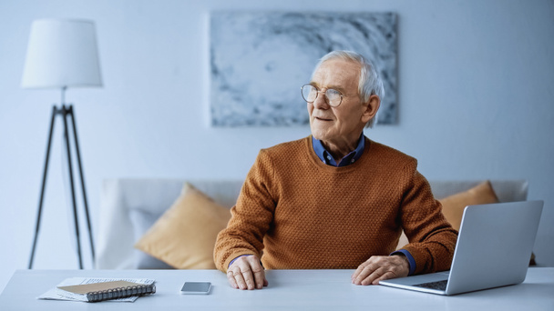 ηλικιωμένος άνδρας κάθεται στο τραπέζι με φορητό υπολογιστή και smartphone στο σπίτι - Φωτογραφία, εικόνα