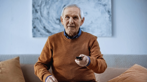 fokussierter älterer Mann sitzt auf Sofa mit Fernbedienung und schaut zu Hause in die Kamera - Foto, Bild