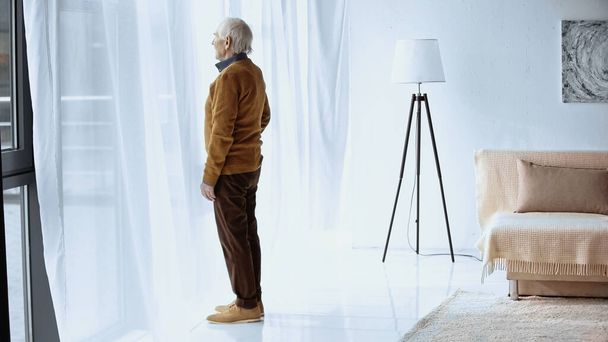 vista de perfil del anciano de pie cerca de la ventana en la sala de estar moderna - Foto, imagen