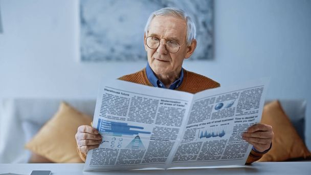 oudere man met een bril die krant leest in de moderne woonkamer - Foto, afbeelding