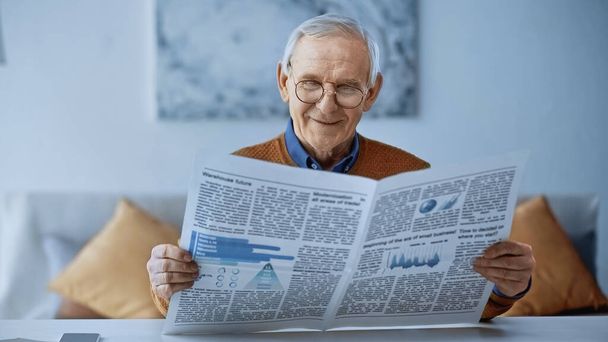 улыбающийся пожилой мужчина в очках, читающий дома газету - Фото, изображение