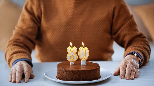 részleges kilátás idős férfi ül előtt születésnapi torta gyertyákkal otthon - Fotó, kép