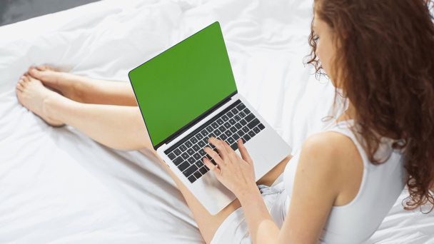 high angle view of curly young woman sitting on bed and using laptop with green screen - Foto, Bild