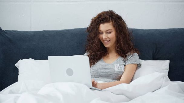 happy freelancer smiling while using laptop in bed  - 写真・画像