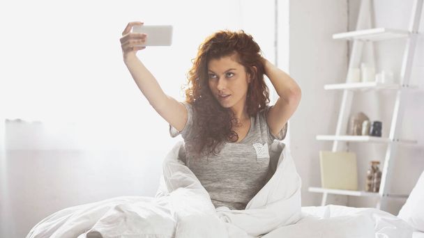Lockige junge Frau macht Selfie mit Smartphone im Schlafzimmer  - Foto, Bild