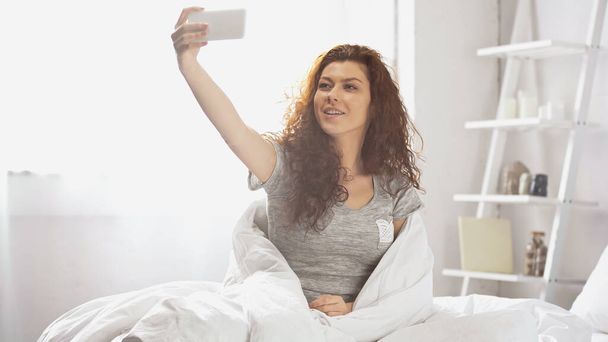 Ευτυχισμένη νεαρή γυναίκα που παίρνει selfie στο smartphone στο υπνοδωμάτιο  - Φωτογραφία, εικόνα