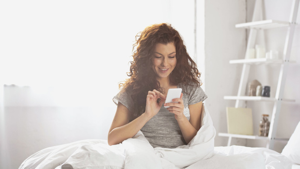 positive young woman messaging on smartphone in bedroom  - Valokuva, kuva
