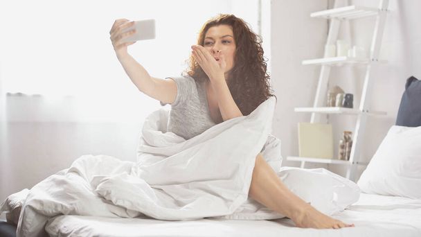 positive young woman sending air kiss while taking selfie on smartphone in bedroom  - Photo, Image