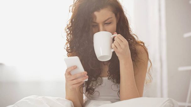 young curly woman drinking coffee and using smartphone  - 写真・画像