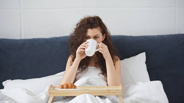 νεαρή γυναίκα πίνει καφέ κοντά στο δίσκο με κρουασάν στο κρεβάτι - Φωτογραφία, εικόνα