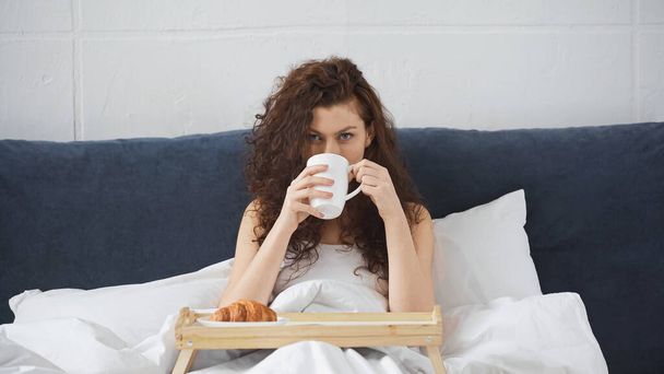 curly woman drinking coffee near tray with croissant on bed - Foto, afbeelding