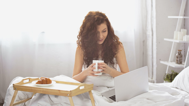šťastná a kudrnatá žena drží šálek kávy a dívá se na notebook v blízkosti tácu s croissant na posteli - Fotografie, Obrázek