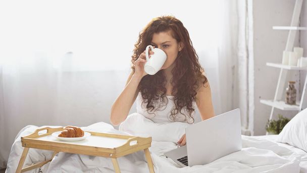 curly young woman drinking coffee near tray with tasty croissant and laptop on bed - Photo, Image