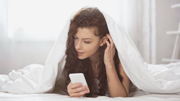 encaracolado jovem mulher usando smartphone enquanto deitado sob cobertor  - Foto, Imagem