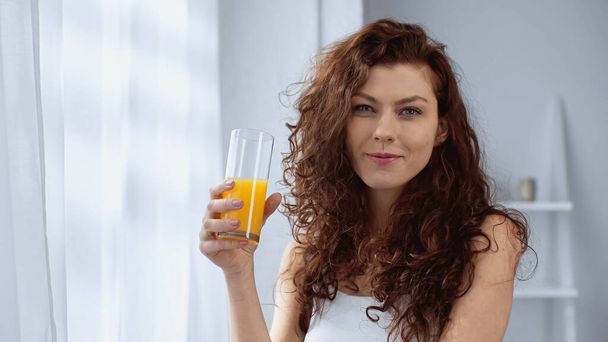 rizado joven mujer sosteniendo vaso de jugo de naranja en casa - Foto, Imagen