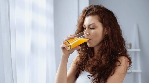 kręcone młoda kobieta trzyma szkło i pije sok pomarańczowy w domu - Zdjęcie, obraz