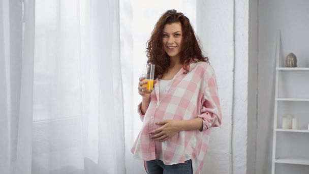 Schwangere und lockige Frau lächelt während sie ein Glas Orangensaft in der Hand hält  - Foto, Bild