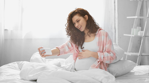 hymyilevä ja raskaana oleva nainen ottaa selfie makuuhuoneessa  - Valokuva, kuva