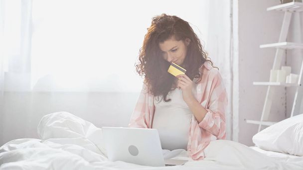 szczęśliwy ciąży młoda kobieta trzyma kartę kredytową podczas korzystania z laptopa w łóżku  - Zdjęcie, obraz