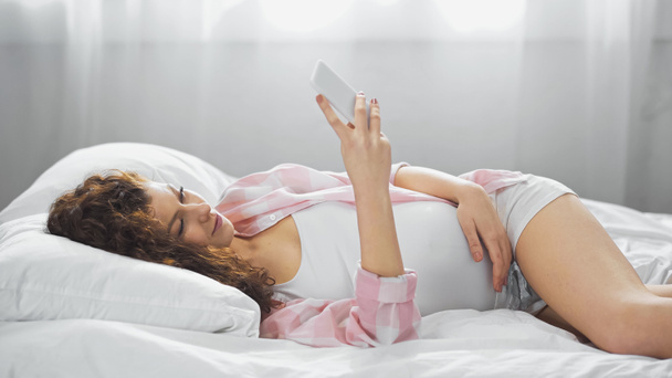 Genç hamile kadın yatakta akıllı telefon kullanıyor. - Fotoğraf, Görsel