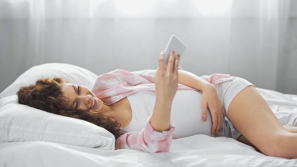 jovem grávida sorrindo enquanto olha para o smartphone na cama - Foto, Imagem