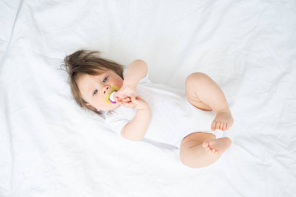 ボディスーツを着た乳首を持つ面白い赤ちゃんが笑顔で家の白い寝具に寝そべってる。トップビュー - 写真・画像