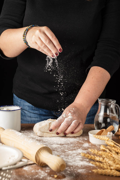 Vista de las manos de la mujer amasando con harina cayendo, sobre mesa de madera con harina y utensilios de cocina, enfoque selectivo, fondo negro, vertical - Foto, Imagen