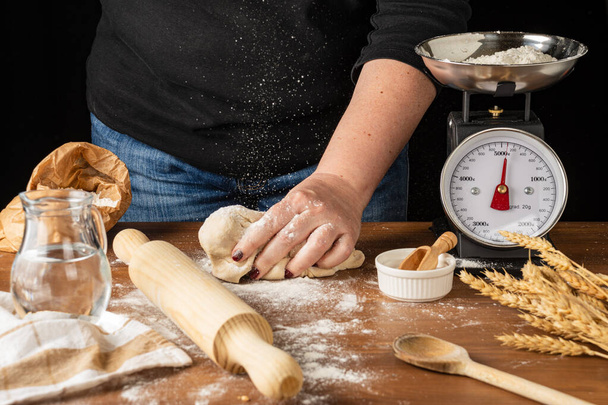 View of woman's hand on pizza dough, falling flour, on wooden table with rustic kitchen objects, black background, horizontal - Photo, Image