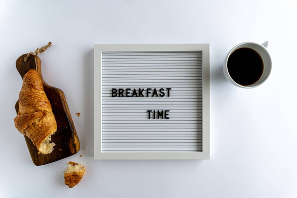 Cartón con palabras que deletrean la hora del desayuno, con un croissant desmenuzable y una taza de café, sobre un fondo blanco, horizontal - Foto, Imagen