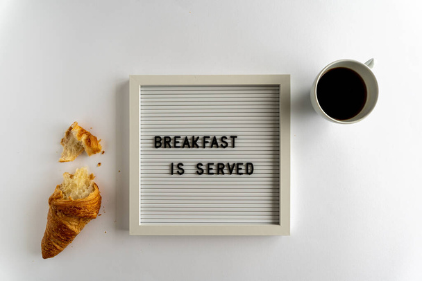 Επιτραπέζιο με λέξεις που συλλαβίζουν το πρωινό σερβίρεται με ένα κρουασάν και ένα φλιτζάνι καφέ, σε λευκό φόντο, οριζόντια - Φωτογραφία, εικόνα