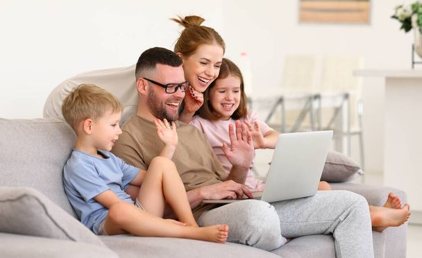 Gyönyörű pár gyerekekkel, fiatal apával, anyával és két gyerekkel, akik videóhívást folytatnak a nagyszülőkkel, integetnek a webkamerának a laptopon és mosolyognak, miközben együtt pihennek otthon a kanapén. - Fotó, kép