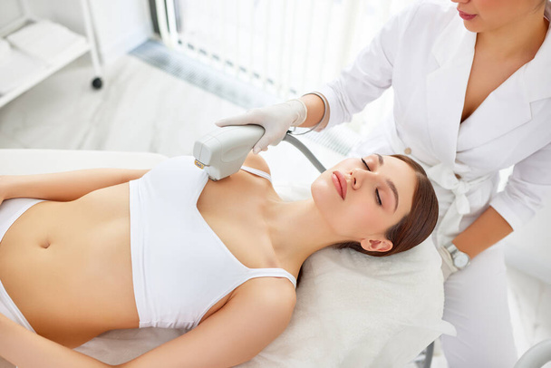 Сверху молодая клиентка получает лазерную процедуру ультразвукового лифтинга кожи во время омоложения в клинике красоты на шее, декольте и груди в современном салоне - Фото, изображение