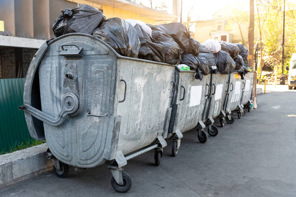 Ряды из многих больших металлических перегруженных мусорных баков, полных черных пластиковых мусорных мешков возле жилого дома в центре города или в пригороде. Неперерабатываемый сортировочный сбор мусора - Фото, изображение